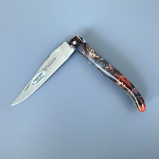 A folding pocket knife with a satin-finished silver blade and a marbled handle featuring black, red, and brown tones with subtle gold flecks, displayed on a solid blue-gray background.