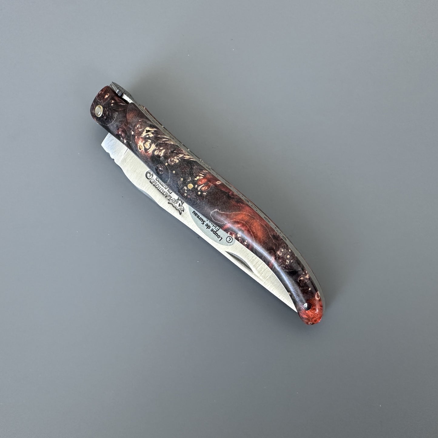 A closed folding pocket knife with a textured, marbled handle in black, red, and brown, and a silver blade, shown on a plain gray background.