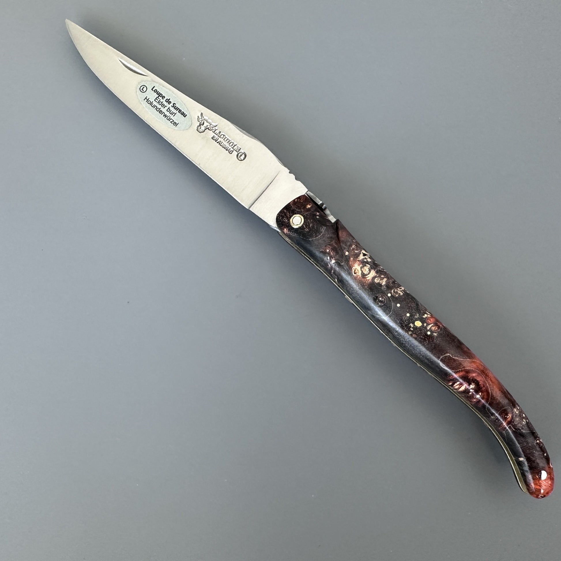 A folding pocket knife with a satin-finished silver blade and a dark brown handle featuring a marbled pattern of black, brown, and subtle red/orange tones. The blade is engraved with the brand name Laguiole en Aubrac