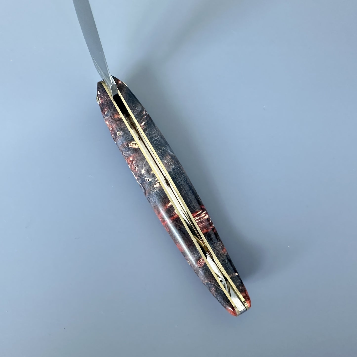 An inner spine of a handcrafted knife handle made of layered brown, orange, and gold-toned materials, shown with a silver blade partially unscrewed at the top against a solid blue-gray background.