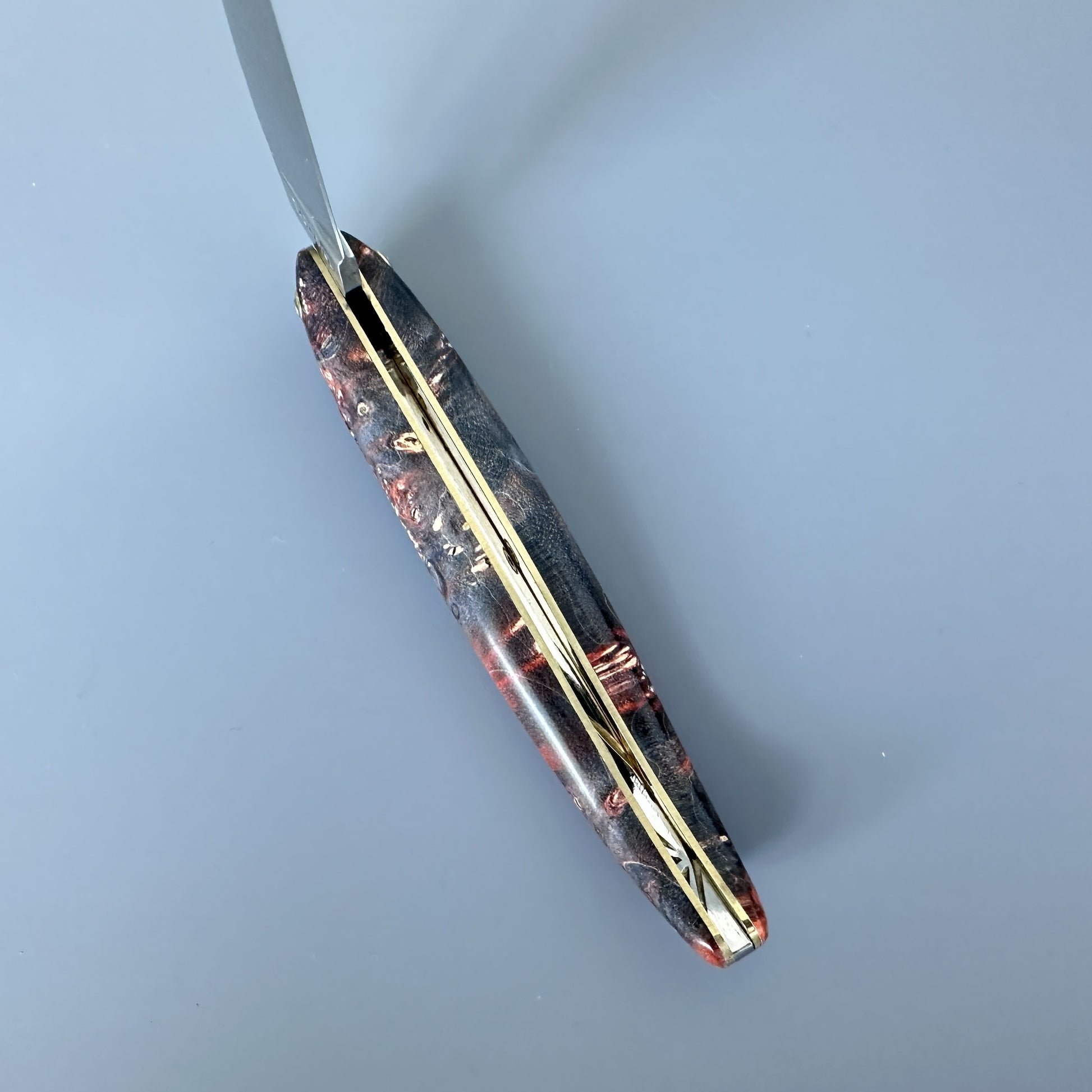 An inner spine of a handcrafted knife handle made of layered brown, orange, and gold-toned materials, shown with a silver blade partially unscrewed at the top against a solid blue-gray background.