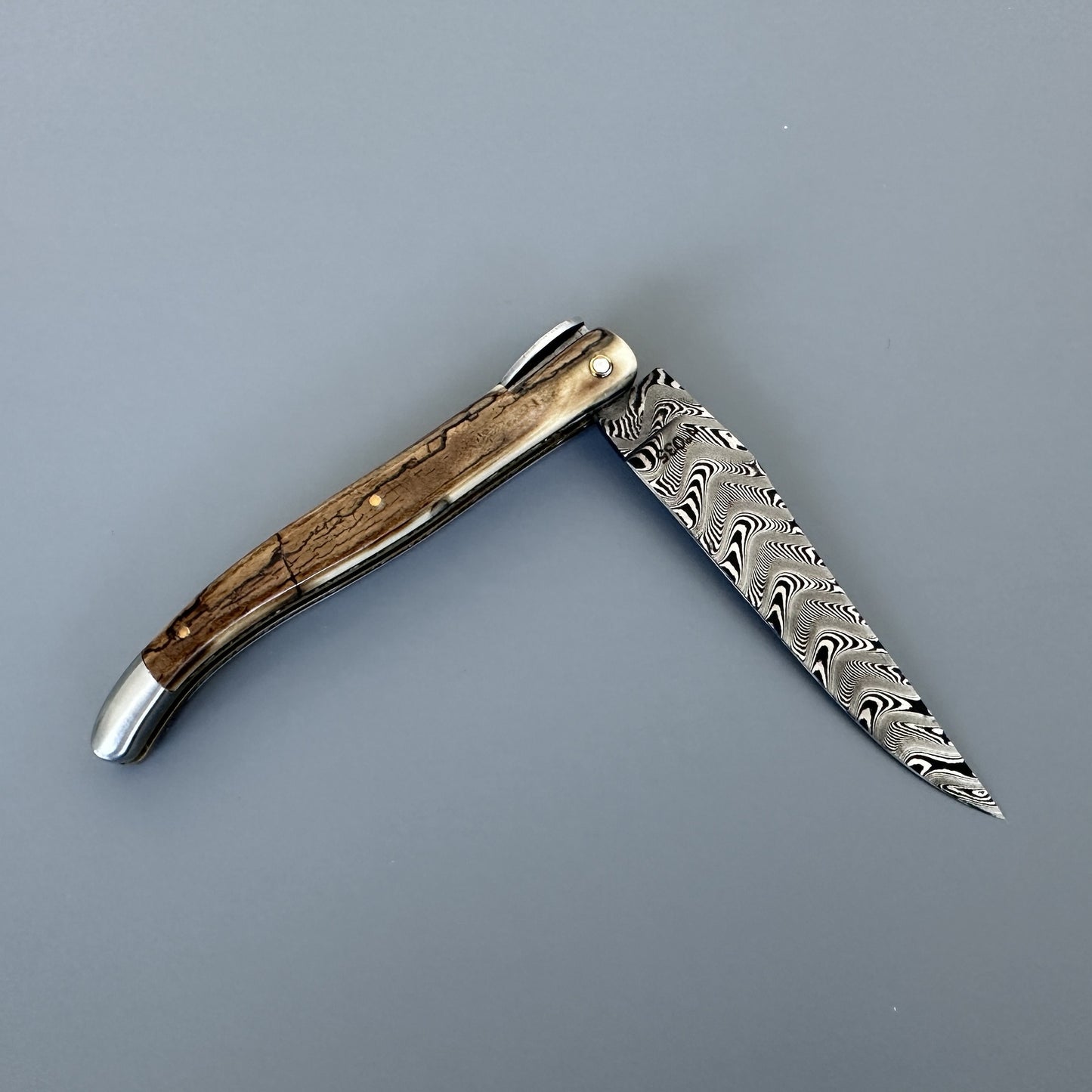 A folding pocket knife with a Damascus steel blade and a curved ivory crust handle, shown open against a solid gray background.