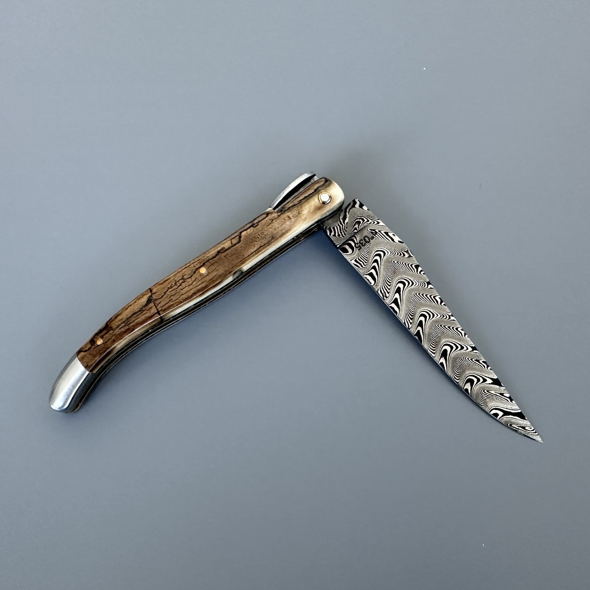 A folding pocket knife with a Damascus steel blade and a curved ivory crust handle, shown open against a solid gray background.