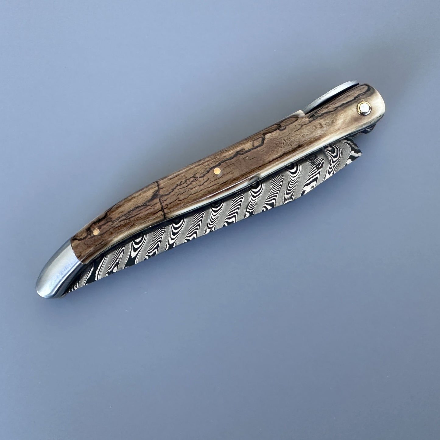 A folding pocket knife with a Damascus steel blade and ivory crust handle, shown on a solid blue-gray background.