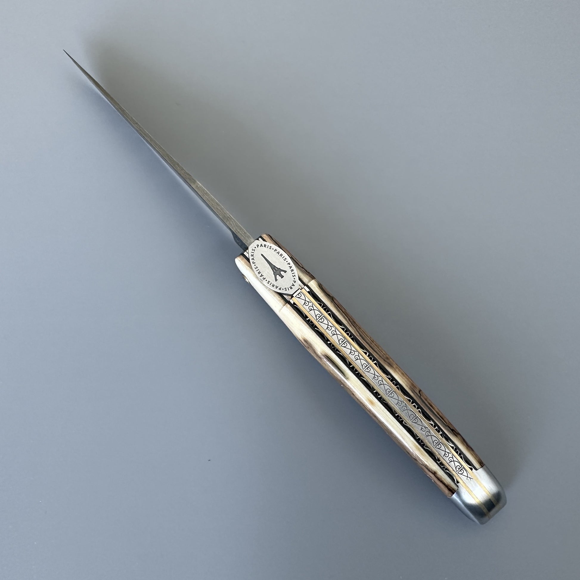 A folding pocket knife with a damascus blade and a ivry crust handle featuring engraved details and a Eifeel tower emblem, displayed on a neutral gray background.