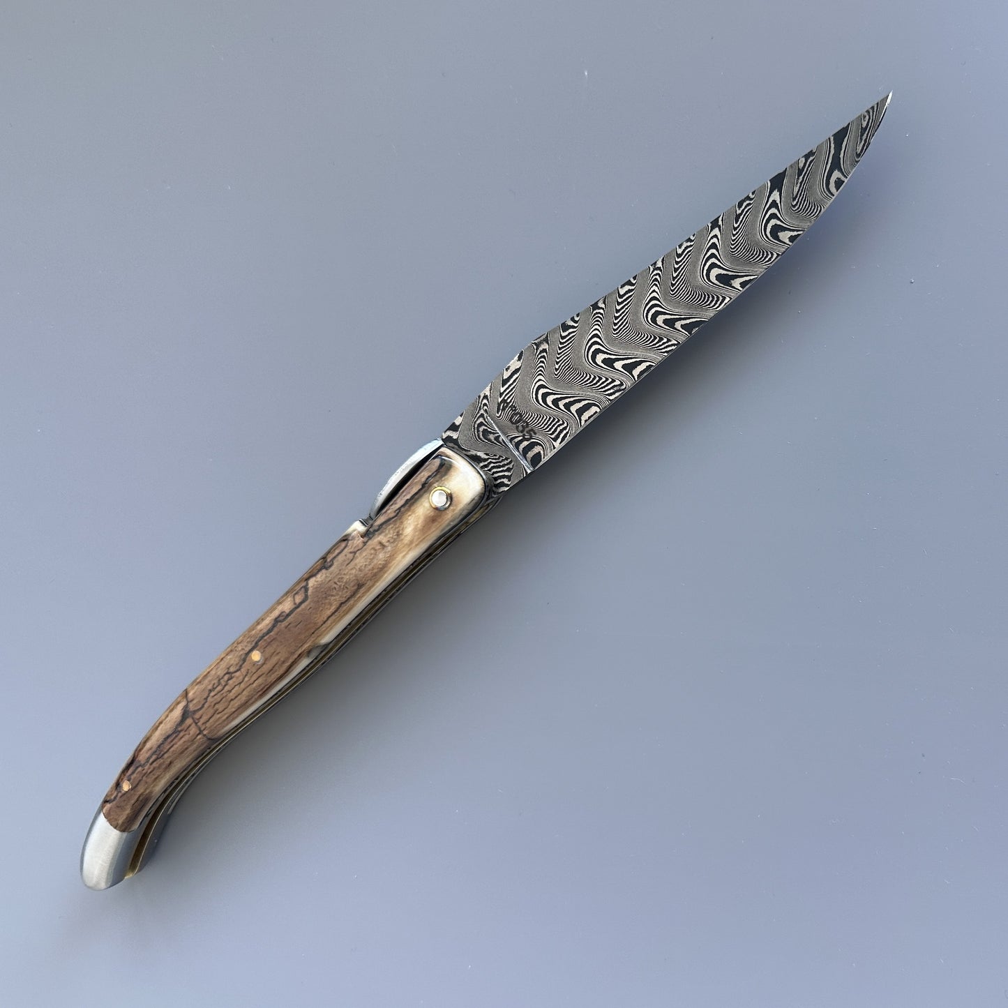A folding pocket knife with a Damascus steel blade and ivory crust handle shown fully open, reverse side.