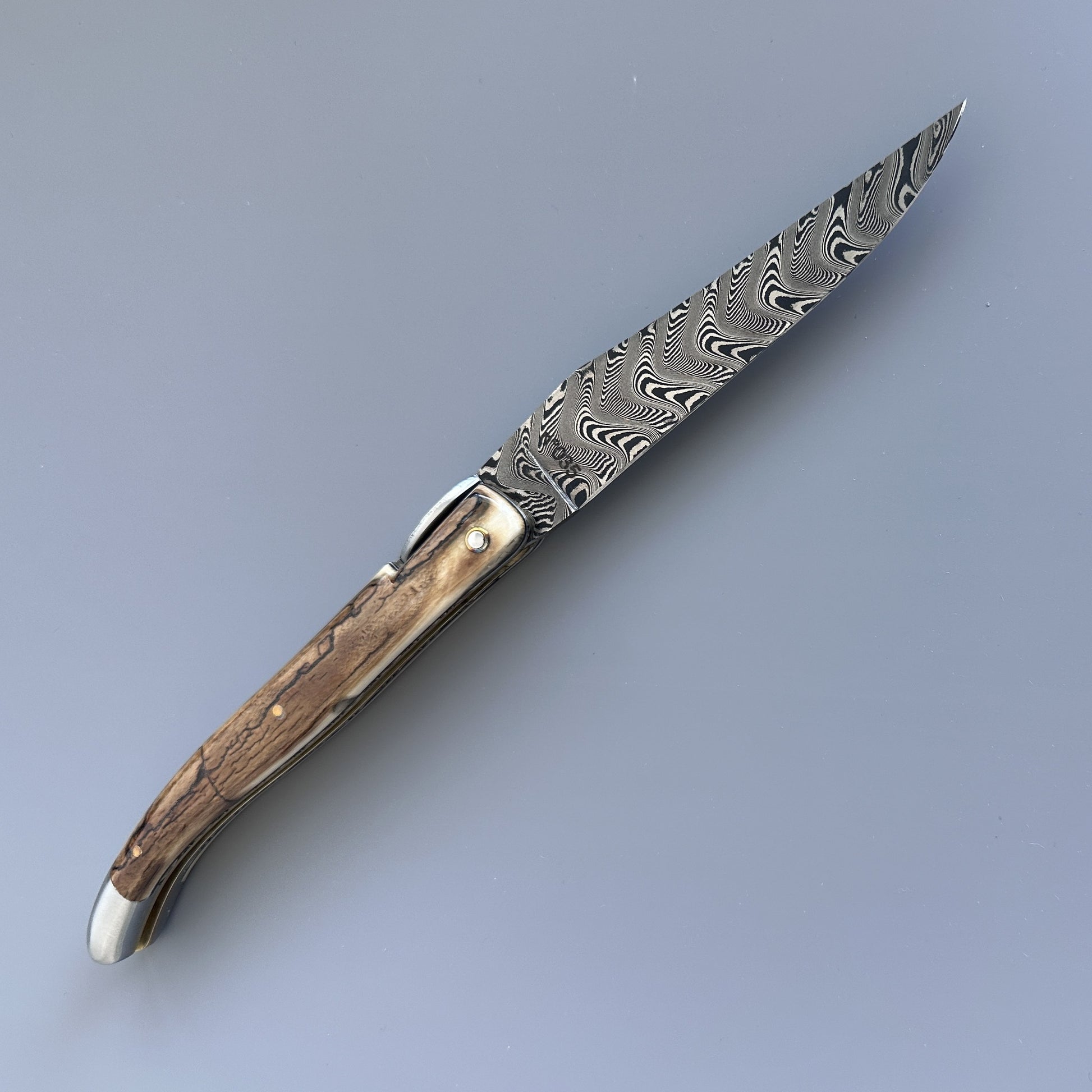 A folding pocket knife with a Damascus steel blade and ivory crust handle shown fully open, reverse side.