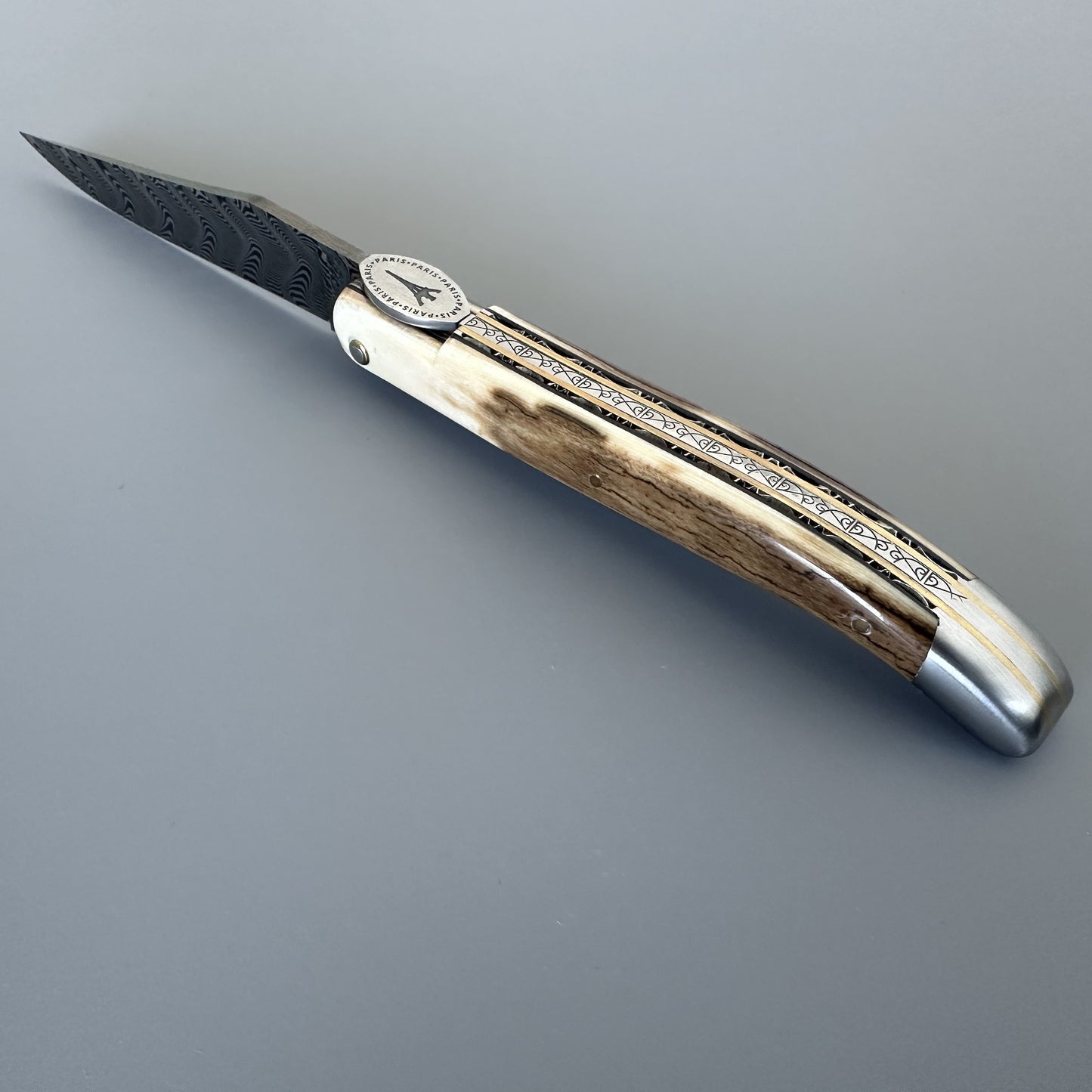 Laguiole en Aubrac folding pocket knife with a Damascus steel blade, featuring a Ivory crust handle with engraved silver and brass inlays and a silver bolster, shown on a neutral gray background.