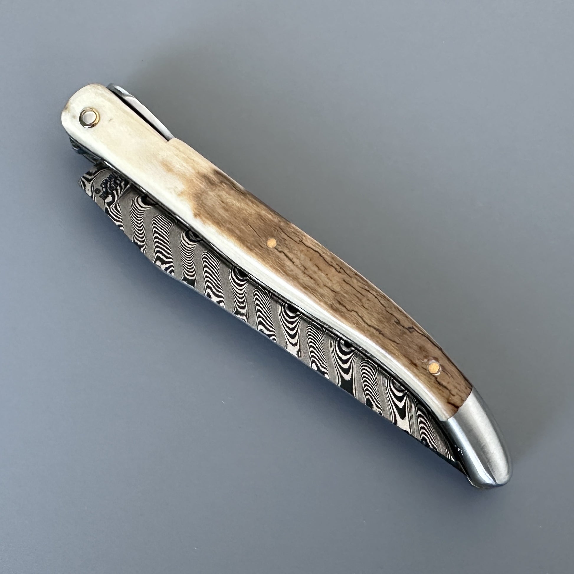 A folding pocket knife with a Damascus steel blade and a handle featuring ivory and natural brown tones in closed position.