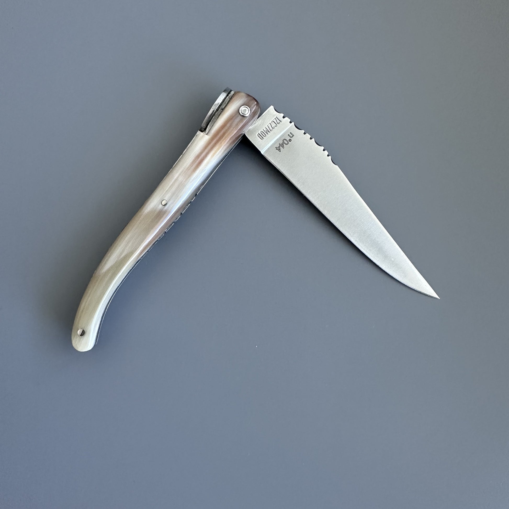A folding pocket knife with a polished silver blade and a curved handle made of natural solid horn, shown open against a solid dark gray background. The blade is engraved with the brand name Laguiole en Aubrac