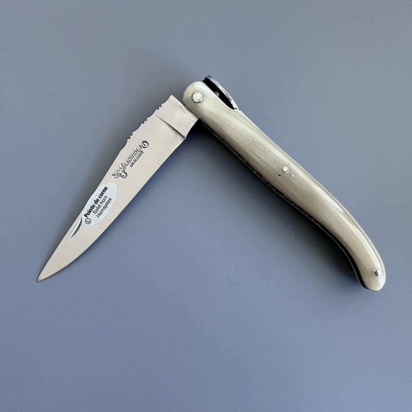 Laguiole en Aubrac folding pocket knife with a silver stainless steel blade and a cream-colored bone handle, shown open on a solid gray background. 