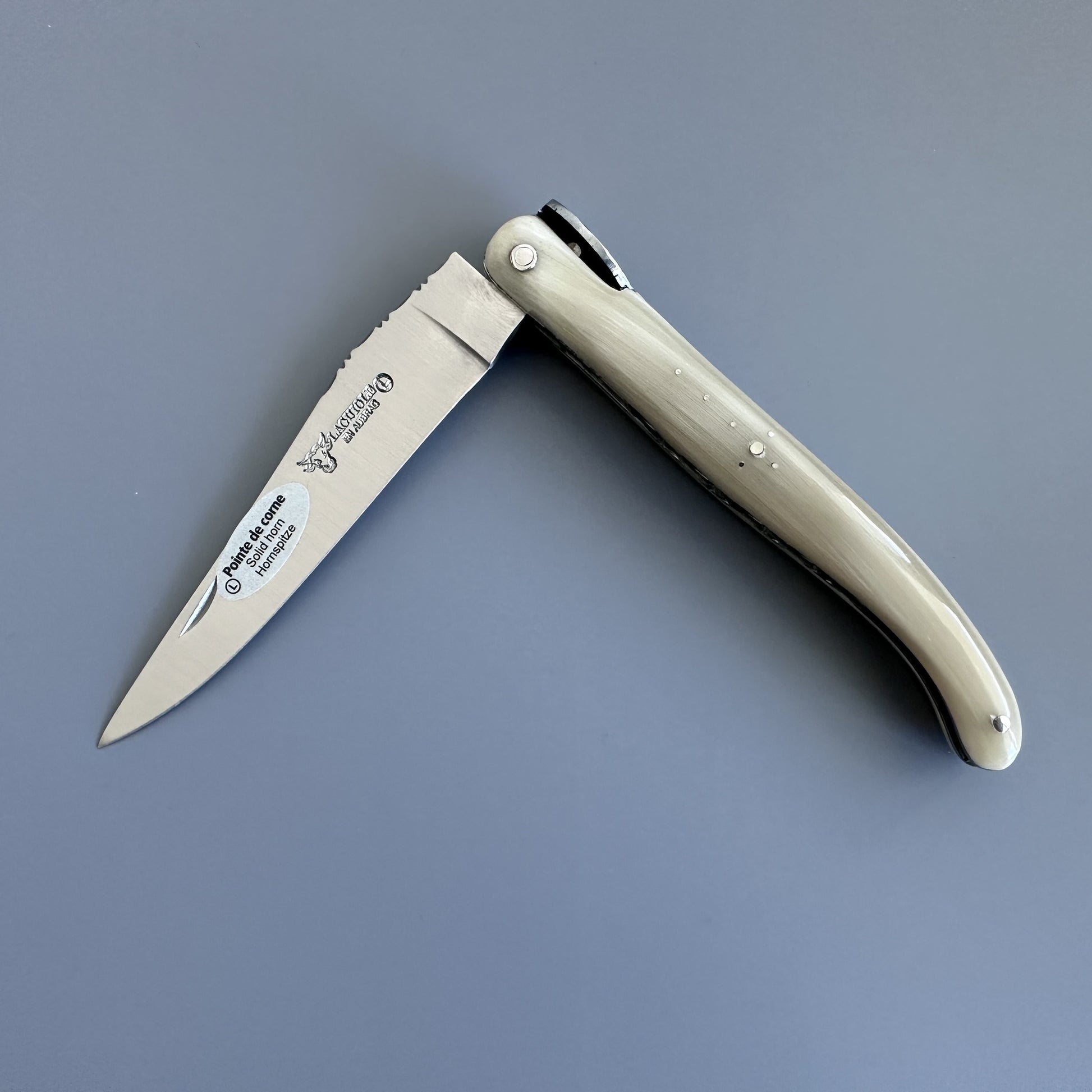 Laguiole en Aubrac folding pocket knife with a silver stainless steel blade and a cream-colored bone handle, shown open on a solid gray background. 