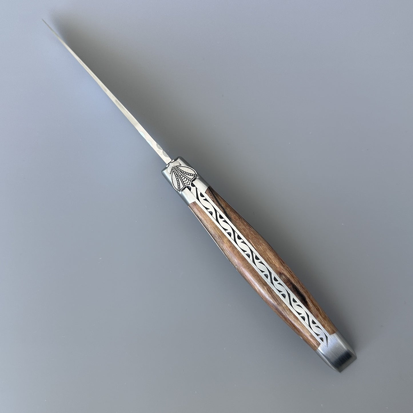 Angled view of the Belle knife's pointed blade and hand-carved bee emblem on the bolster, Aubrac wood handle