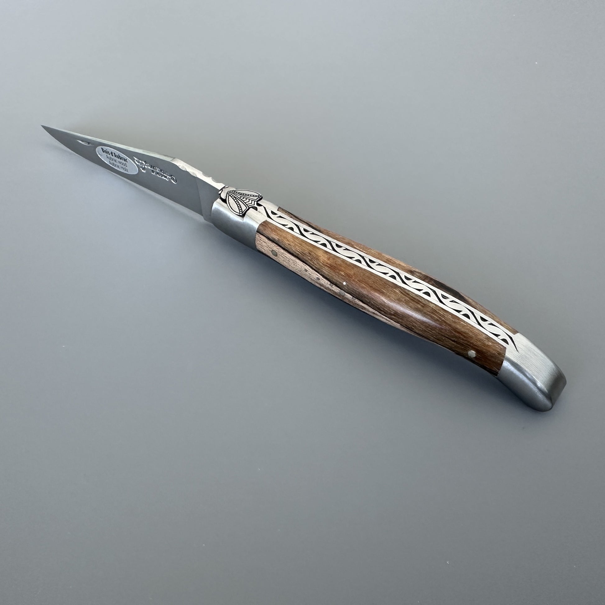 Angled view of the Belle knife's pointed blade and hand-carved bee emblem on the bolster, Aubrac wood handle