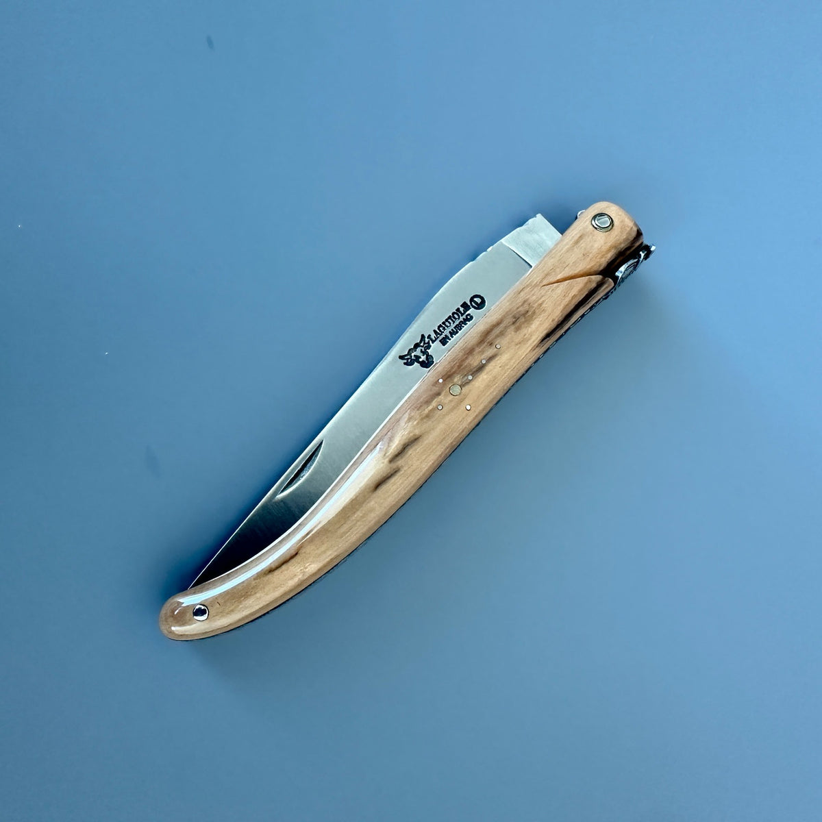 A folding pocket knife with a satin silver blade and a light brown color handle, shown open on a solid blue background. 