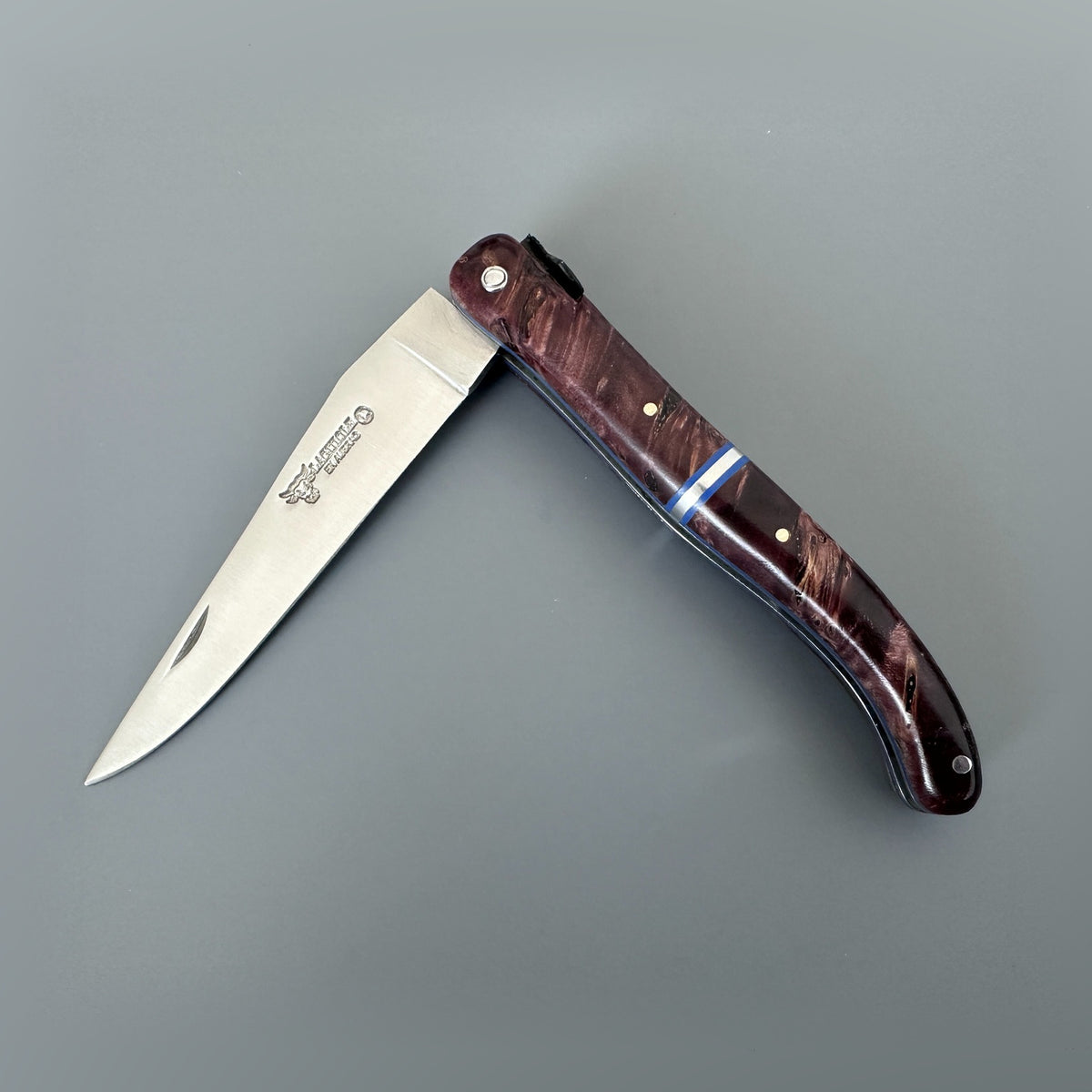 Laguiole en Aubrac folding pocket knife with a mat silver blade and a wine reddish-brown wood-grain handle, shown open on a neutral gray background. The handle features a blue inlay accent and brass pins.