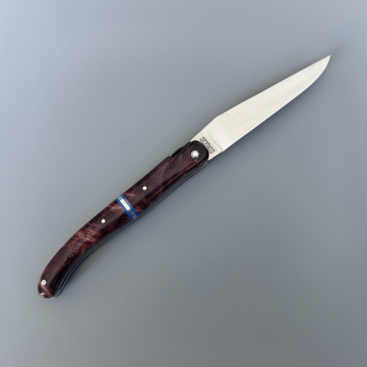 Laguiole en Aubrac fully open knife with a satin blade and wine brown poplar burl handle featuring a blue inlay accent, shown on a neutral gray background.
