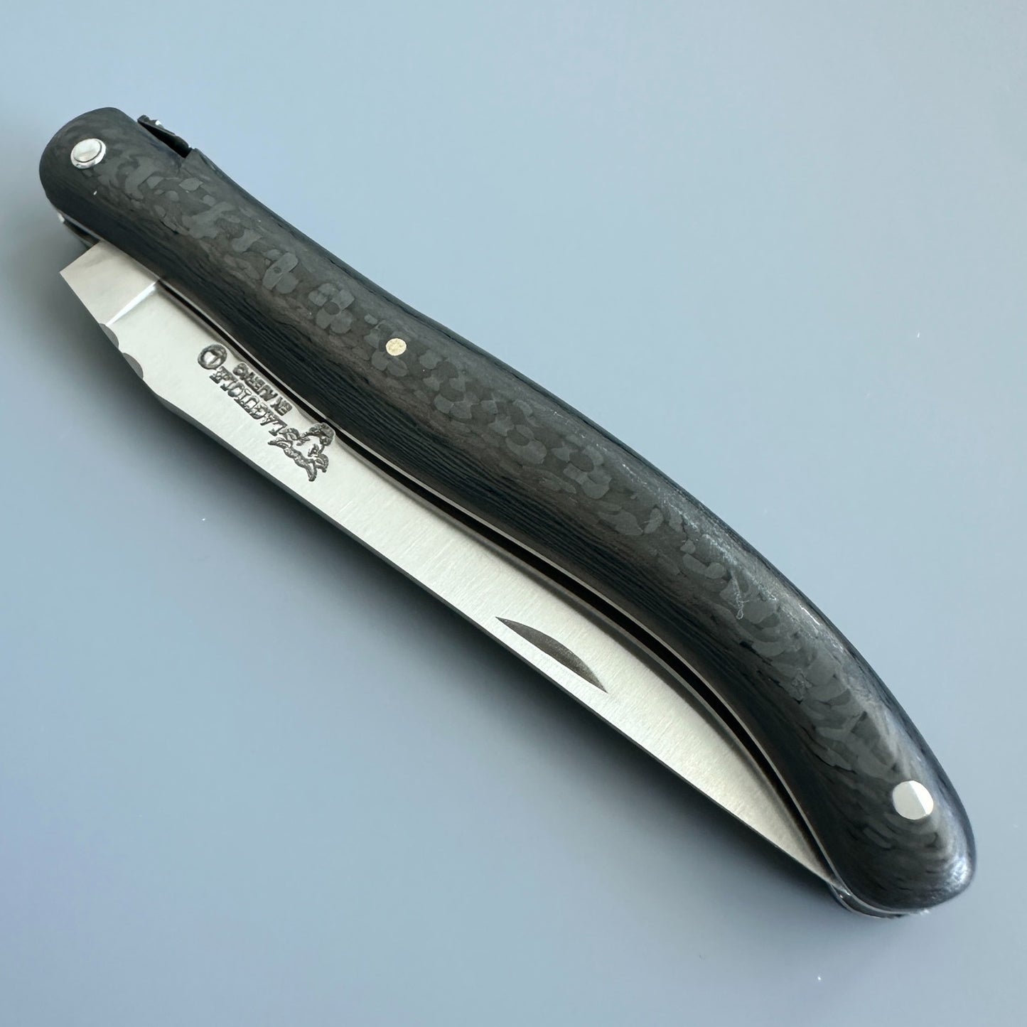 Closed Laguiole en Aubrac Belle Piece Carbon folding knife with the blade logo visible beside its curved carbon handle over blue background