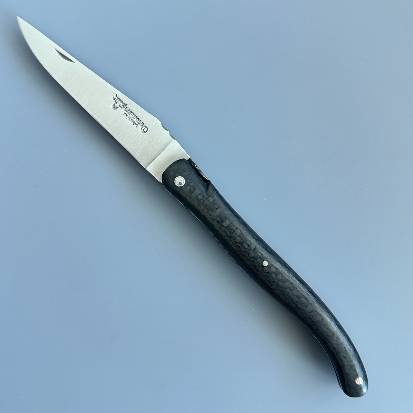 Open Laguiole en Aubrac Belle Piece Carbon folding knife showing its blade, patterned handle, geometric bee and engraved spine.