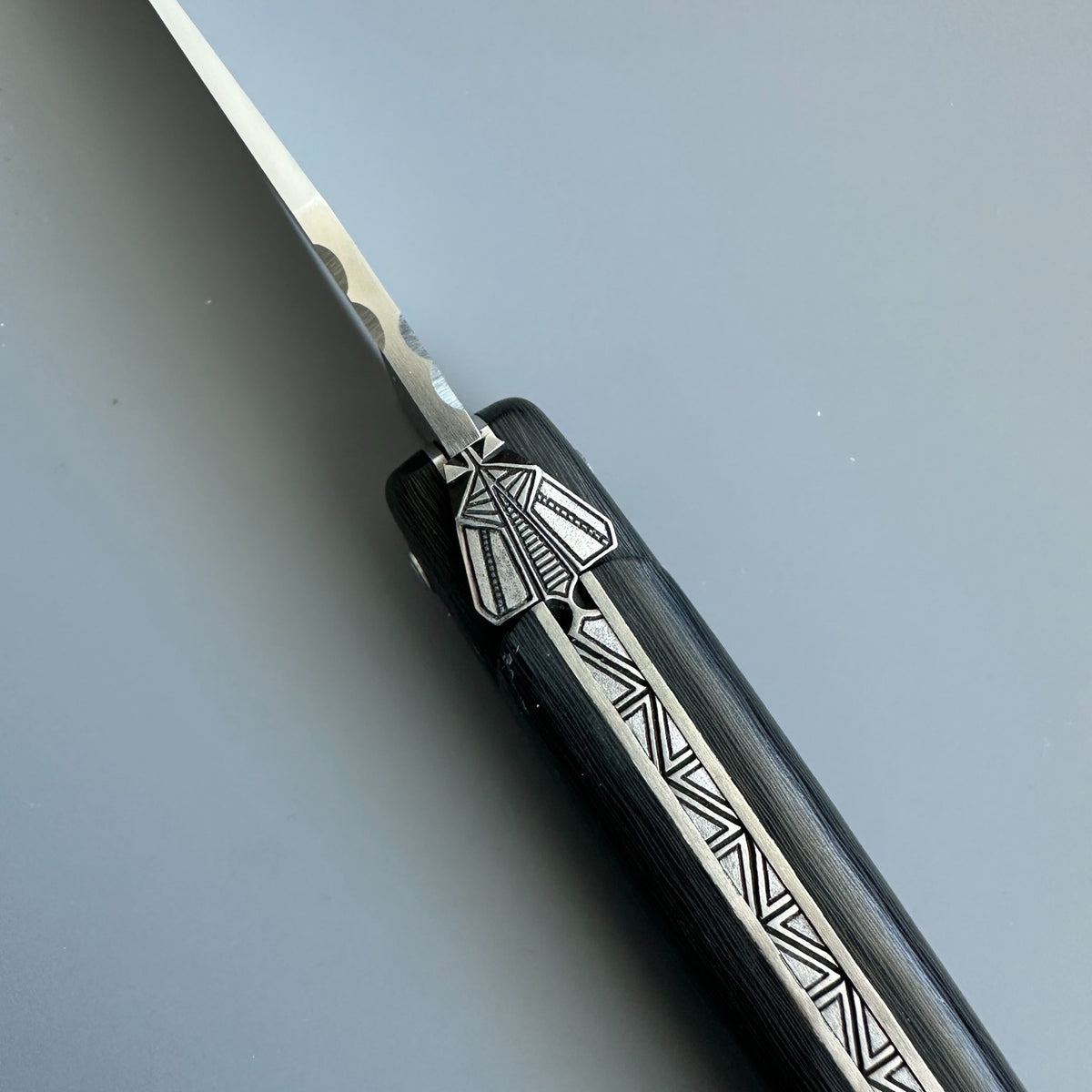Close-up of the geometric bee ornament and engraved spine on a Laguiole en Aubrac Belle Piece Carbon folding knife over blue background