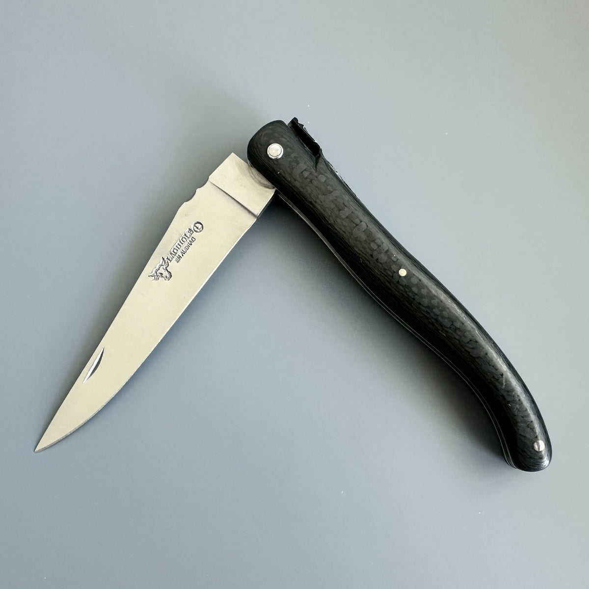 Laguiole en Aubrac Belle Piece Carbon folding knife partly open, showing the blade logo and dark patterned handle.