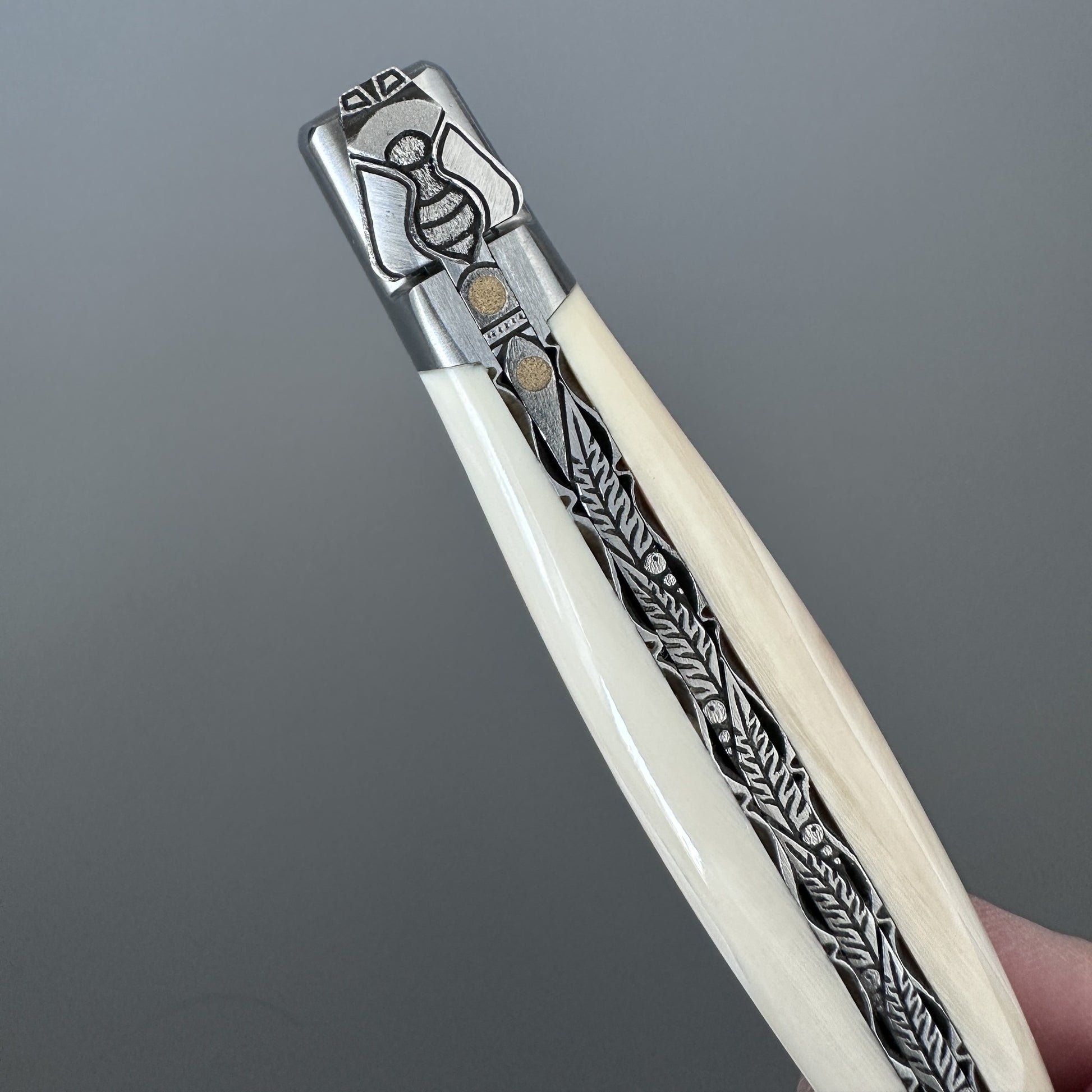 Close-up of the Laguiole en Aubrac Belle knife's bee bolster and fern-leaf engraving on warthog bone handle, hand-held