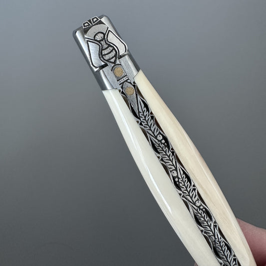 Close-up of the Laguiole en Aubrac Belle knife's bee bolster and fern-leaf engraving on warthog bone handle, hand-held