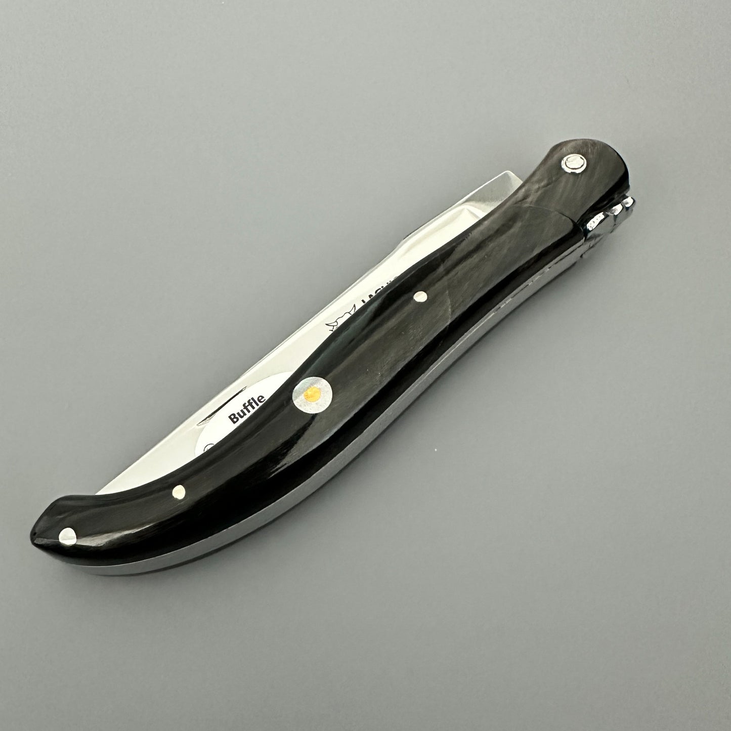Profile view of the closed Laguiole en Aubrac folding knife with its black buffalo-horn handle and decorative emblem over gray background