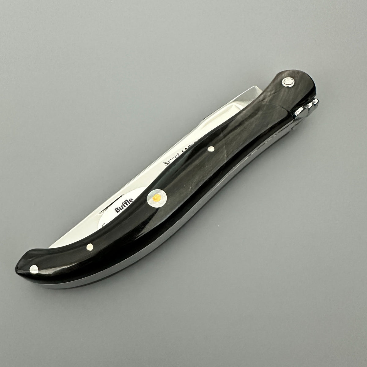 Profile view of the closed Laguiole en Aubrac folding knife with its black buffalo-horn handle and decorative emblem over gray background