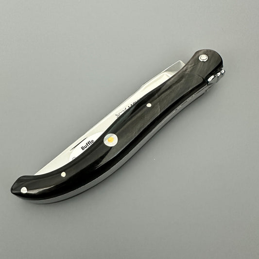 Profile view of the closed Laguiole en Aubrac folding knife with its black buffalo-horn handle and decorative emblem over gray background