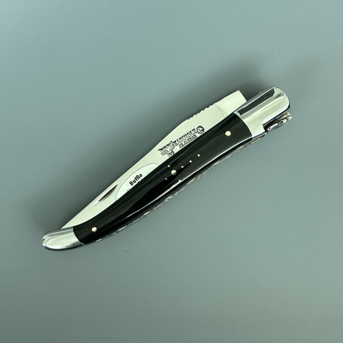A folding pocket knife with a buffalo horn handle and polished silver blade, shown open on a plain gray background. The brand Laguiole en Aubrac engraved in the blade.
