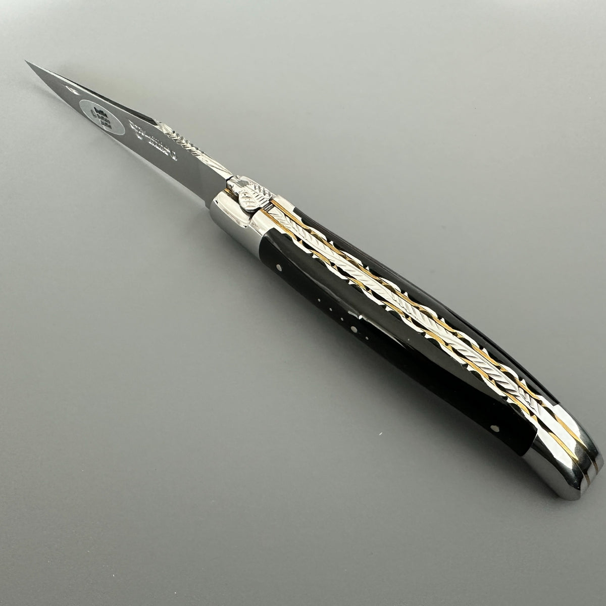 Laguiole en Aubrac folding knife with a buffalo handle featuring intricate brass and silver filigree inlays, and a silver stainless steel blade, shown on a neutral gray background.