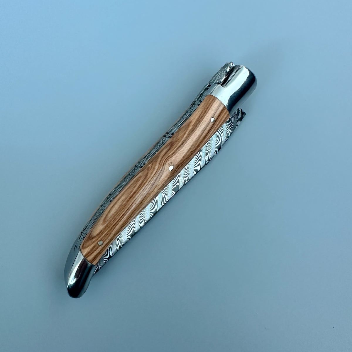 A folding pocket knife with a Damascus steel blade partially drawn, featuring a natural wood handle and polished silver metal accents, shown on a solid light blue background.