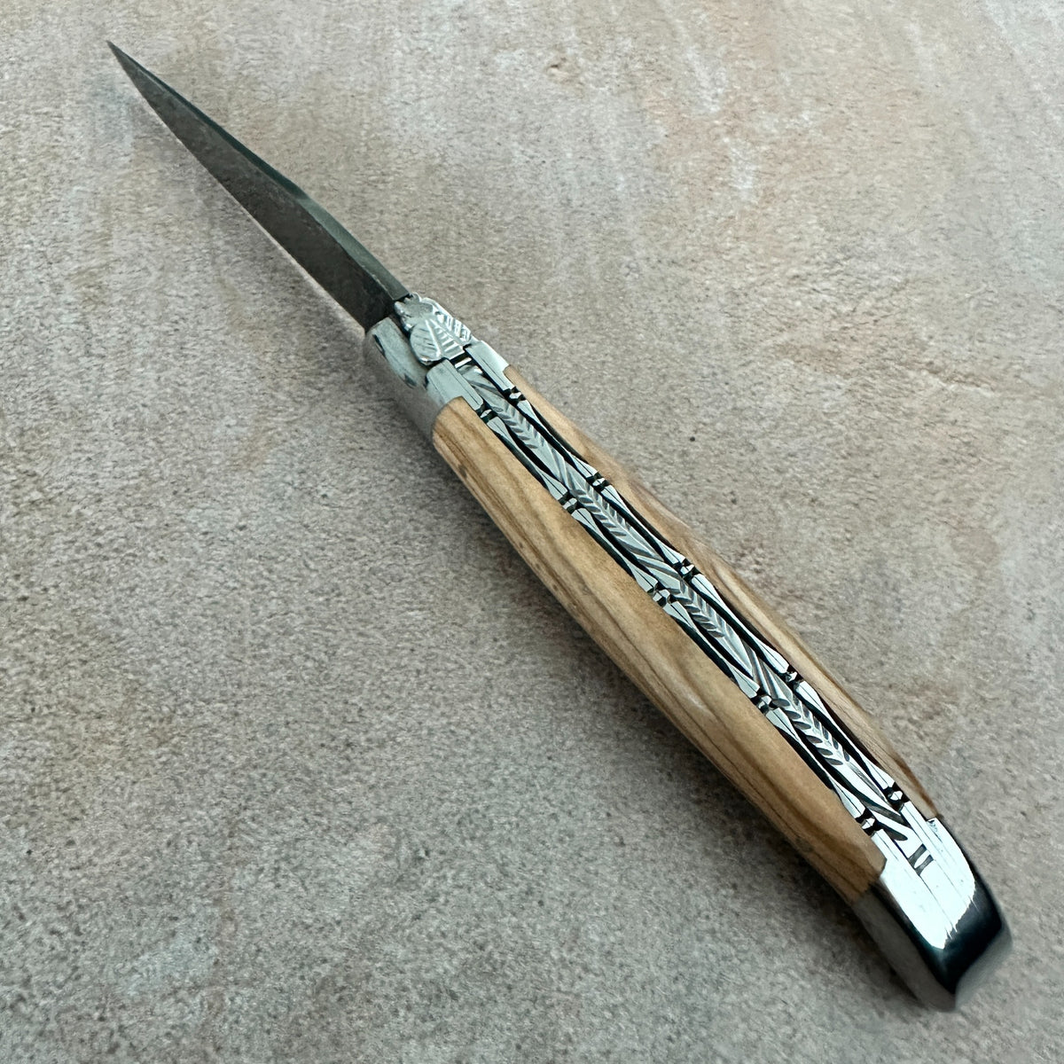 A folding pocket knife with a light brown wood handle featuring black inlay patterns and silver metal accents, resting on a textured beige surface.