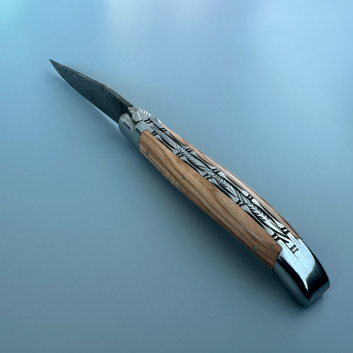 A folding pocket knife with a natural wood handle featuring engraved silver metal details, shown open on a smooth, light blue-gray background.