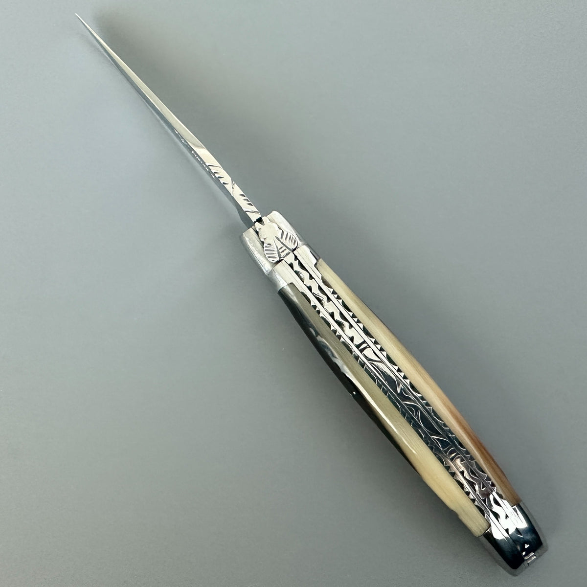 A folding Laguiole en aubrac pocket knife with a polished silver steel blade and solid horn handle, featuring intricate engraved patterns, resting diagonally on a neutral gray background.