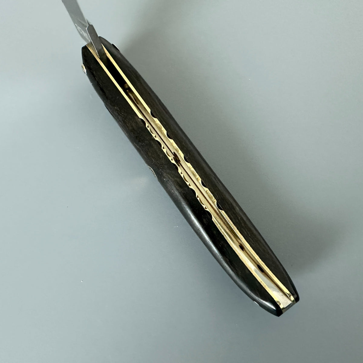 A Laguiole en Aubrac close-up of a folding pocket knife with  Ebony wooden handle and gold brass accents, with the blade partially extended, set against a plain light gray background.