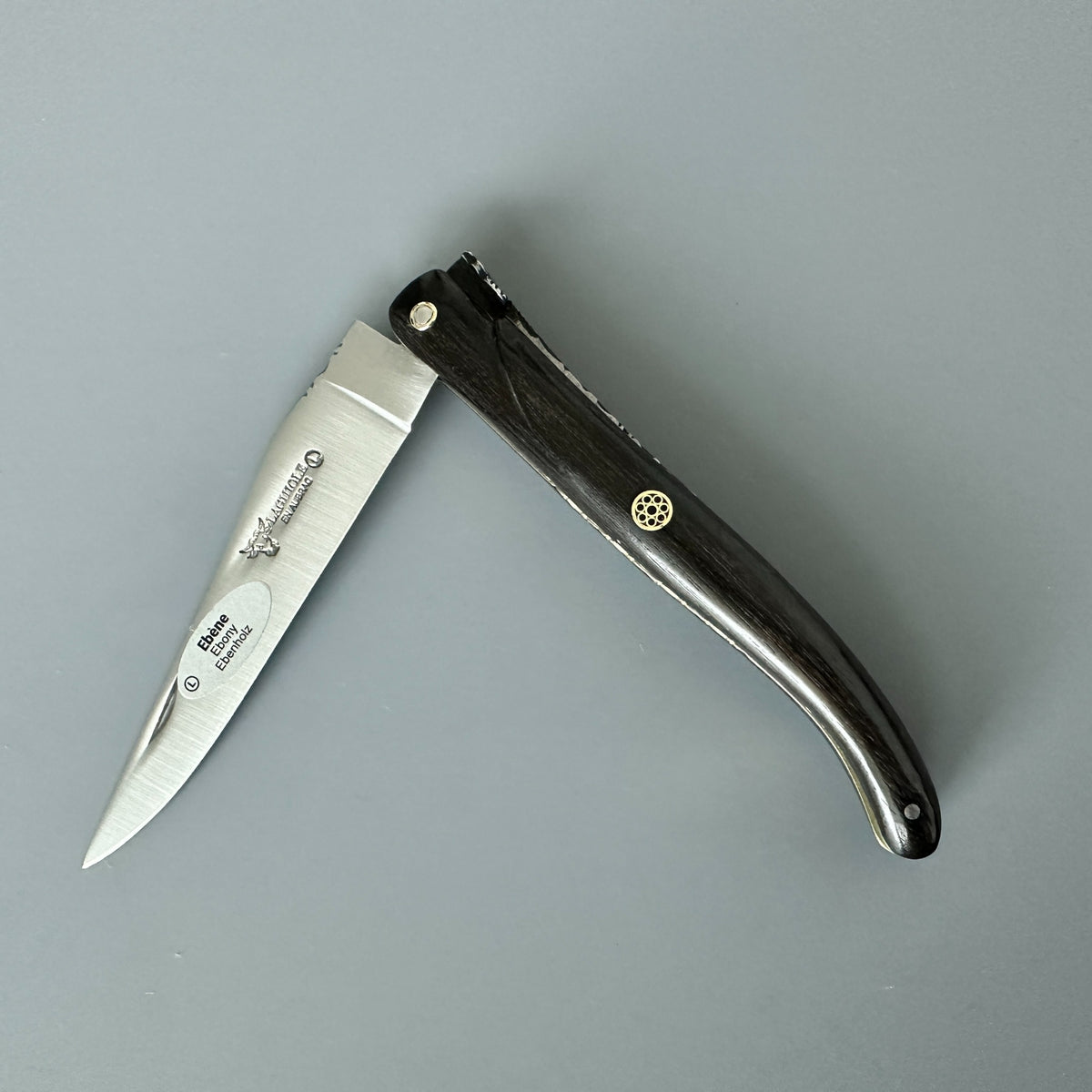 Laguiole en Aubrac folding knife with Ebony handle and brass mosaic pin partially open over a dray background