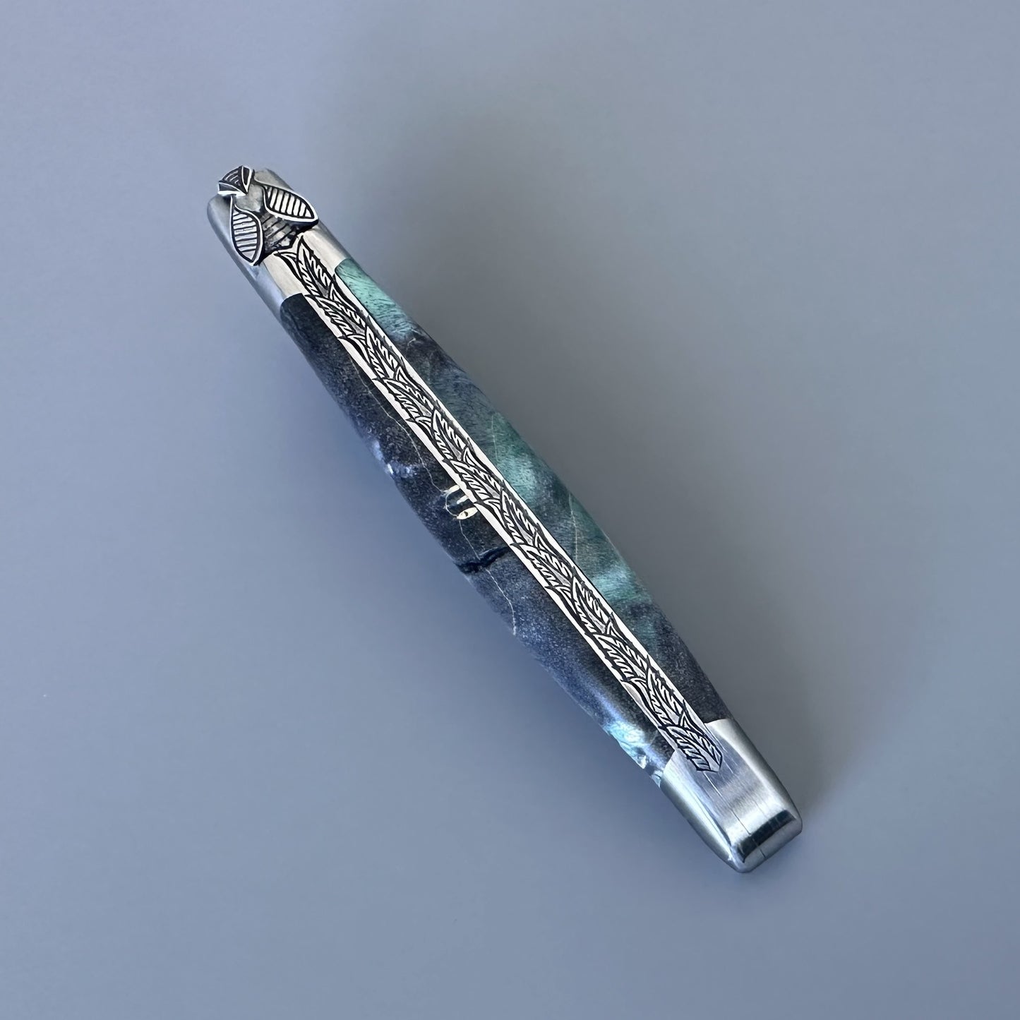 Diagonal close-up of an open Laguiole en Aubrac knife showing the blue-green elder burl scales and engraved fern-leaf bolster detail