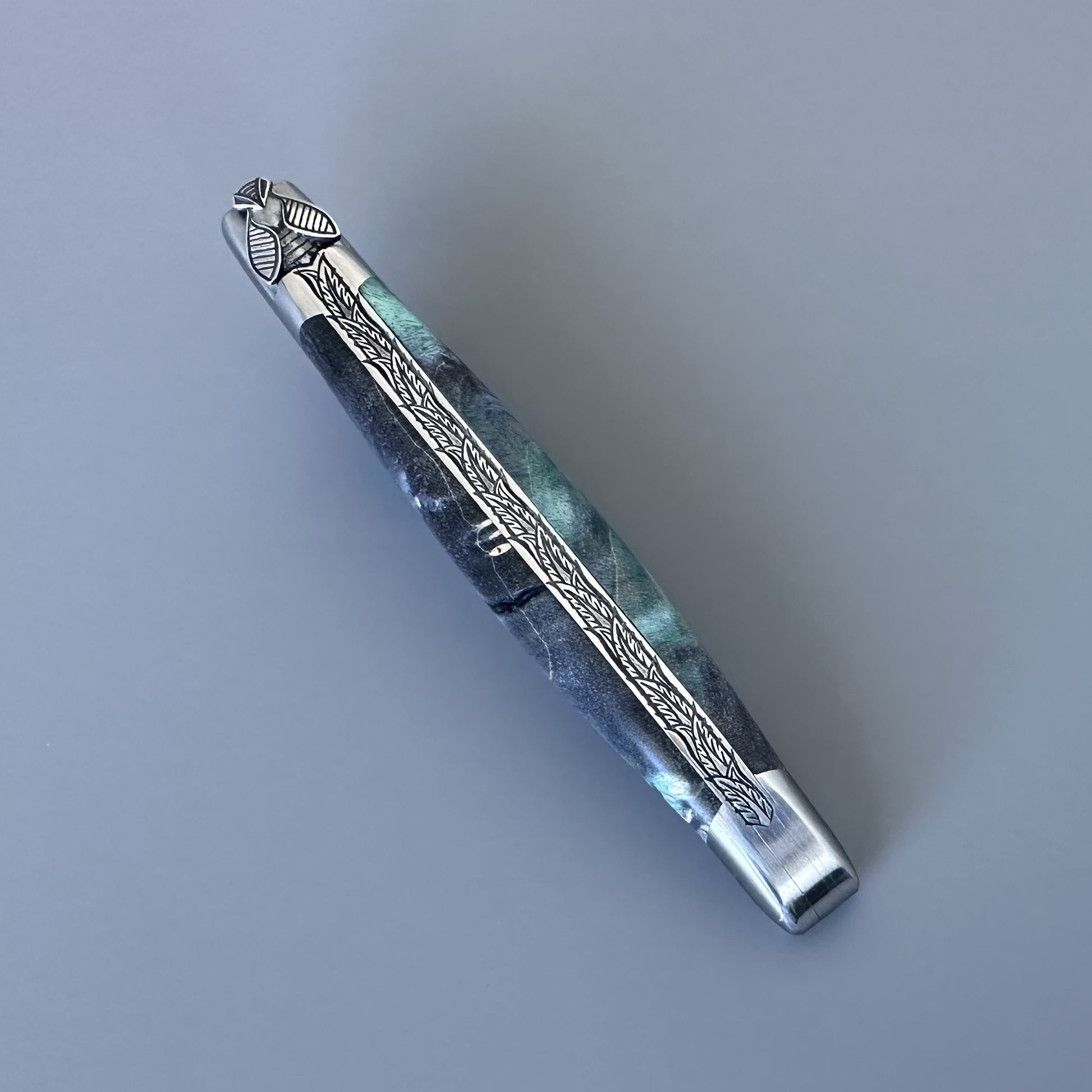 Diagonal close-up of an open Laguiole en Aubrac knife showing the blue-green elder burl scales and engraved fern-leaf bolster detail