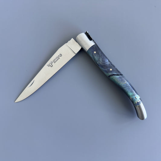 Open Laguiole en Aubrac folding knife with polished steel blade and blue-green elder burl handle featuring the bee emblem, shown blade-left on a gray background