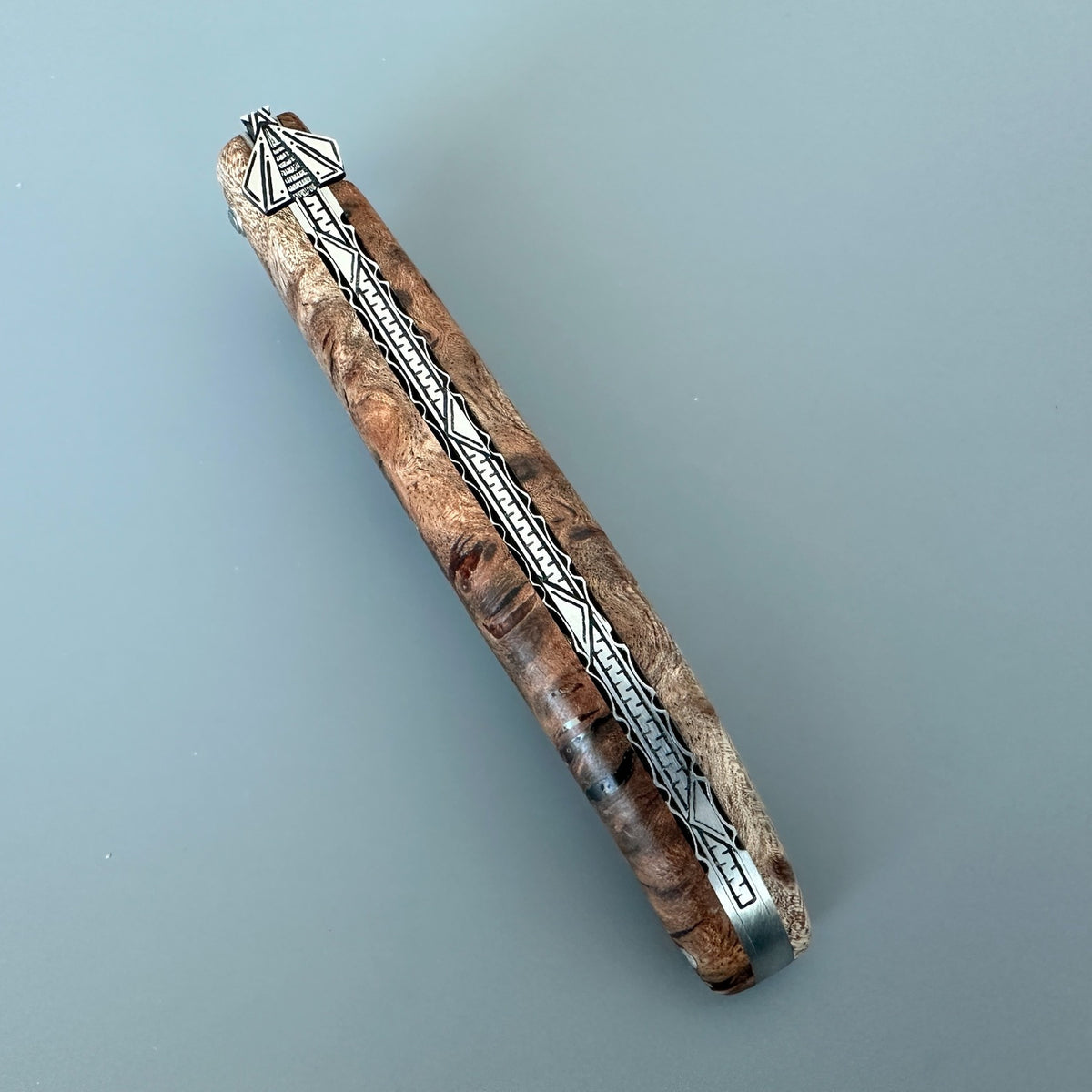 Laguiole en Aubrac Belle Piece Knife with custom engraved Spine showing  a rich Albizia burl wood grain pattern in shades of brown and beige, accented with silver metal geometric engravings and a silver cap.