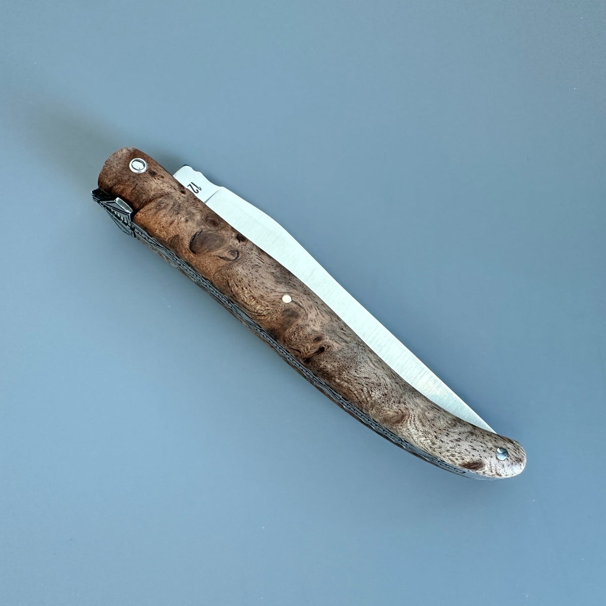 Reverse view of Laguiole en Aubrac folding pocket knife with a textured, Albizia burl wood handle and satin metal blade,  on a solid blue-gray background. 