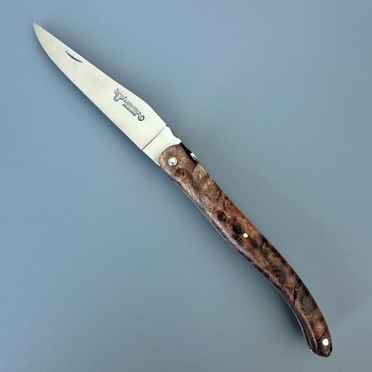 Laguiole en Aubrac Belle Piece with Albizia Burl handle shown diagonally on a solid blue-gray background. 