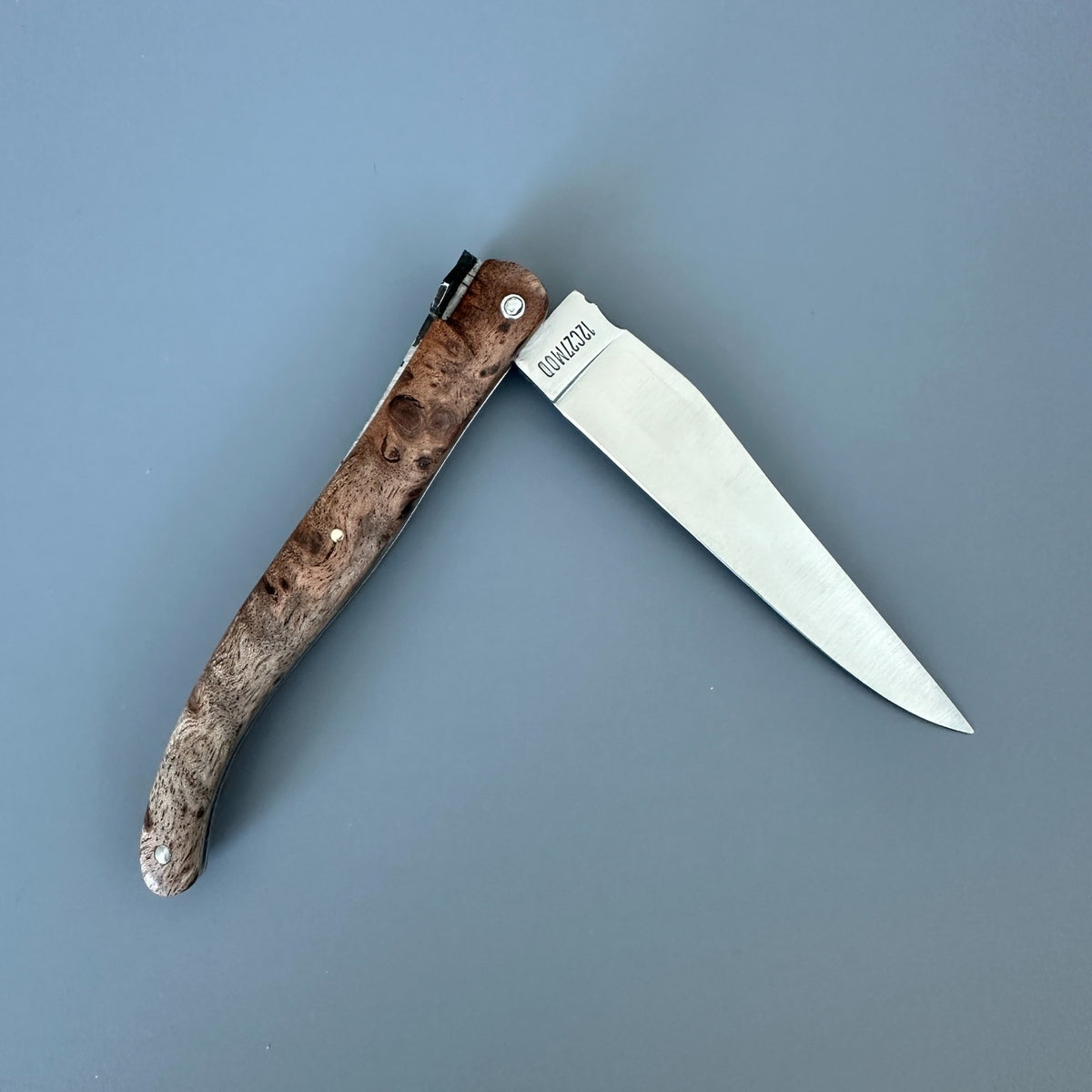 Reverse side of Laguiole en Aubrac Belle Piece folding  knife with a satin blade and Albizia burl wood handle, shown open on a solid slate blue background. 