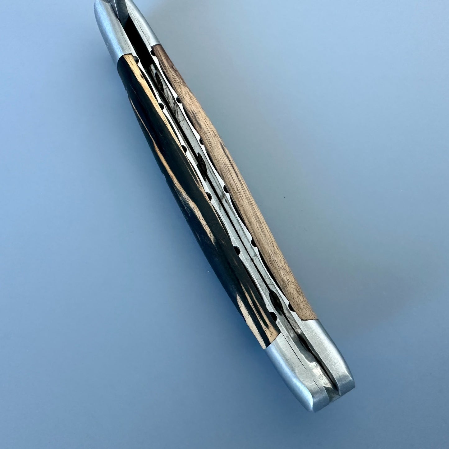 Laguiole en Aubrac Handcarved knife with a silver-toned metal frame, a dark wood inlay, and a black handle with gold-toned stripes, shown open on a light blue background.