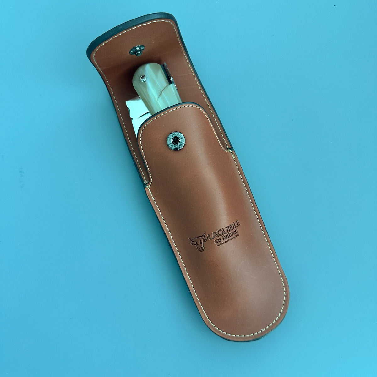 A tan leather knife sheath containing a hunting folding knife with a The sheath features white contrast stitching, a snap button closure, and the brand name and logo: 'Laguiole en Aubrac"