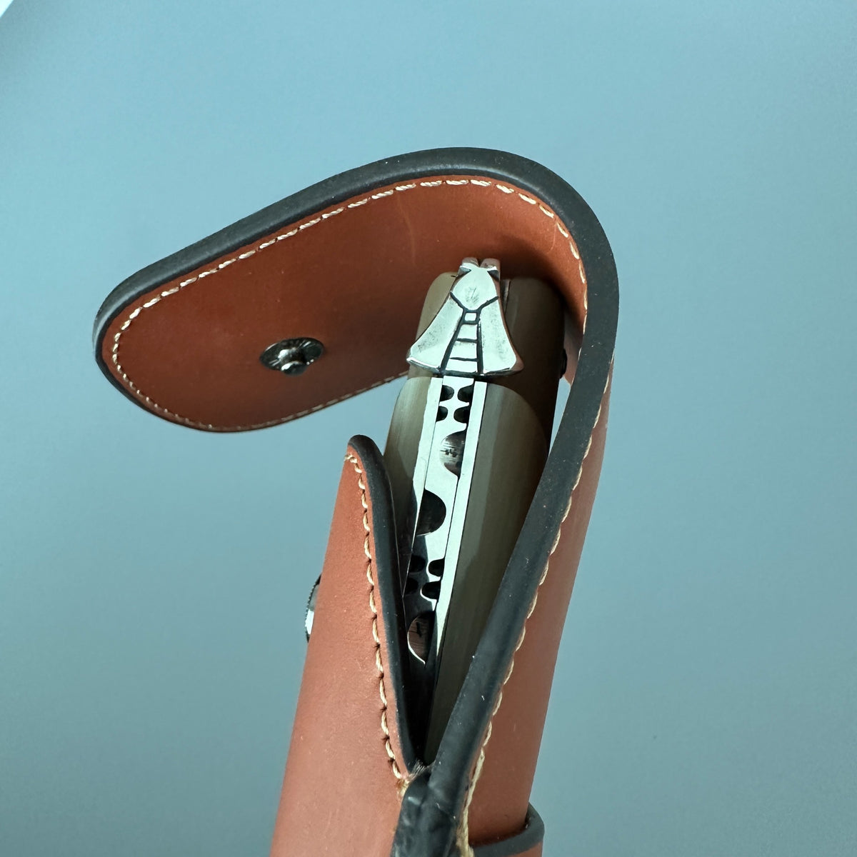 A close-up of a Laguiole en Aubrac Hunting Knife L0514 partially tucked into a brown leather sheath with black trim, set against a smooth light blue background.