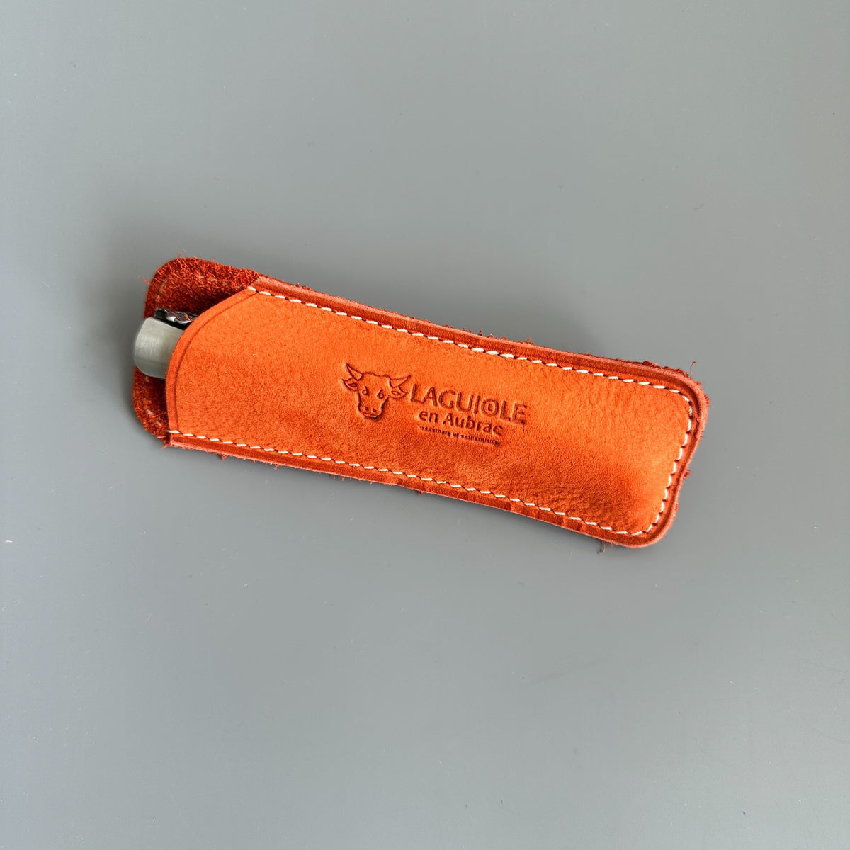 An orange leather pocket lighter case with white stitching, featuring an embossed logo and brand name 'LAGUIOLE en Aubrac' on a neutral gray background.