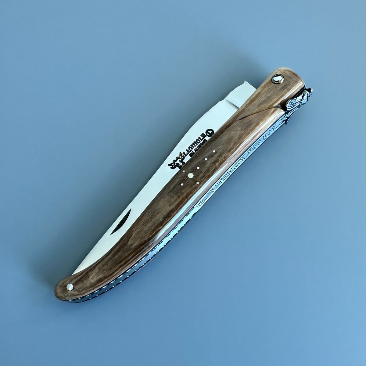 Laguiole en Aubrac mammoth ivory folding knife closed diagonally, showing the handle pins, blade logo and decorated spine